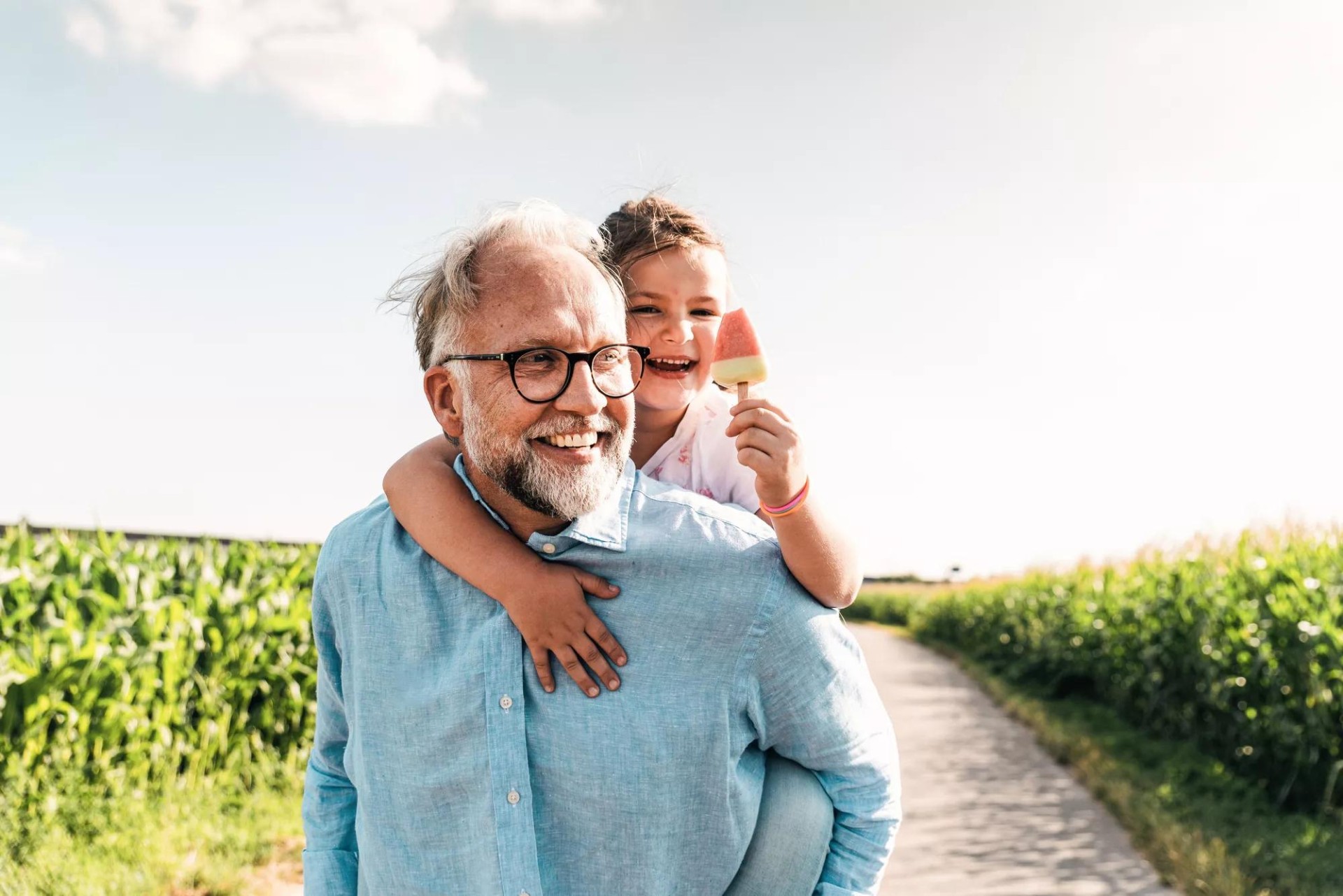 Father joyfully carries his young child on his back through a field, while the child enjoys a popsicle.